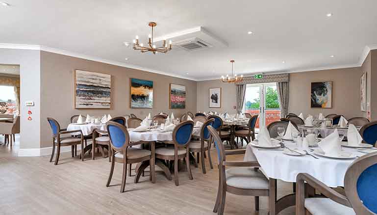 An elegant dining room with multiple set tables, grey and blue upholstered chairs, and paintings on the walls under a coffered ceiling with a chandelier.