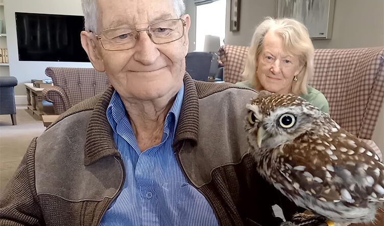 An elderly man holding a small owl on their arm in a living room