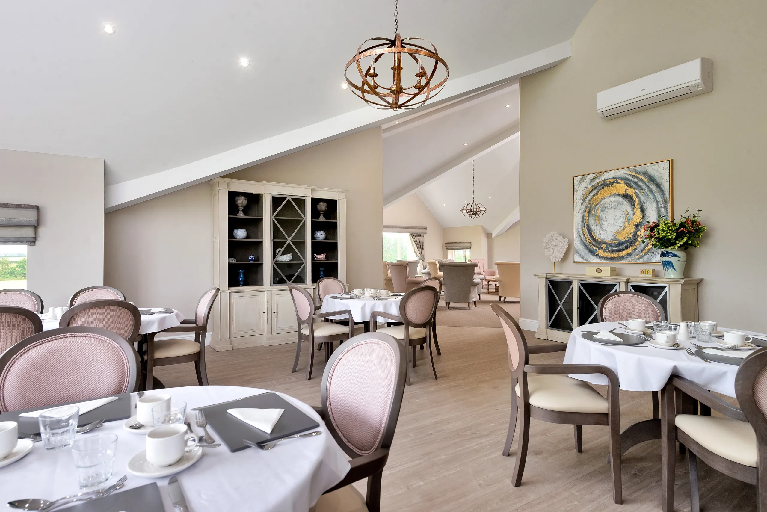 The elegant dining room at Roden Hall Care Home, with tables dressed in white linen, dusky-pink upholstered chairs, a display cabinet and artwork on the walls.