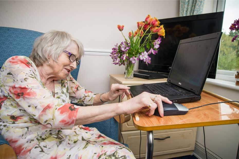 An elderly lady using a laptop at a desk with a bouquet of flowers and a black monitor in the background