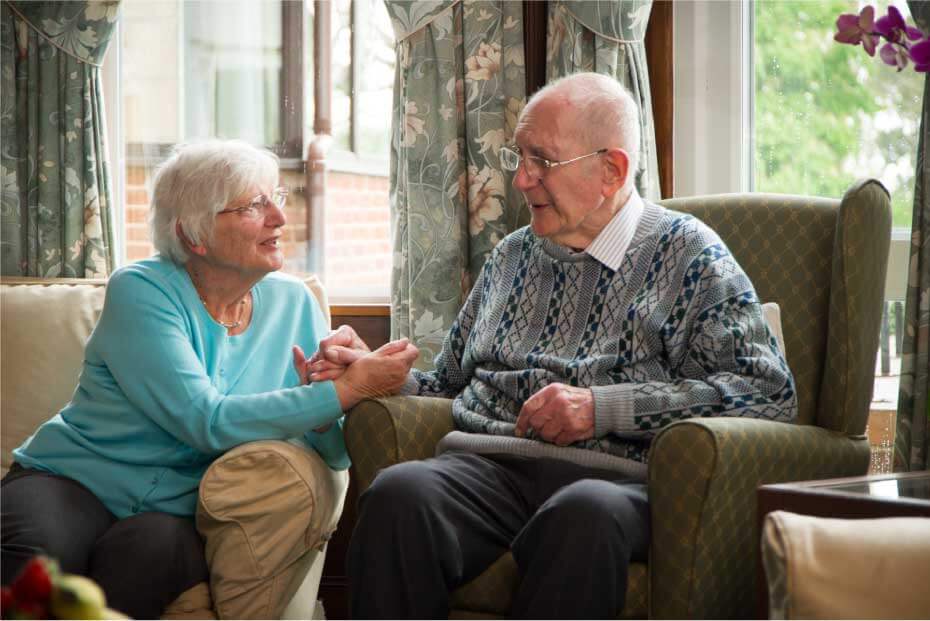 An elderly man and woman sitting on a couch, holding hands and engaging in a conversation