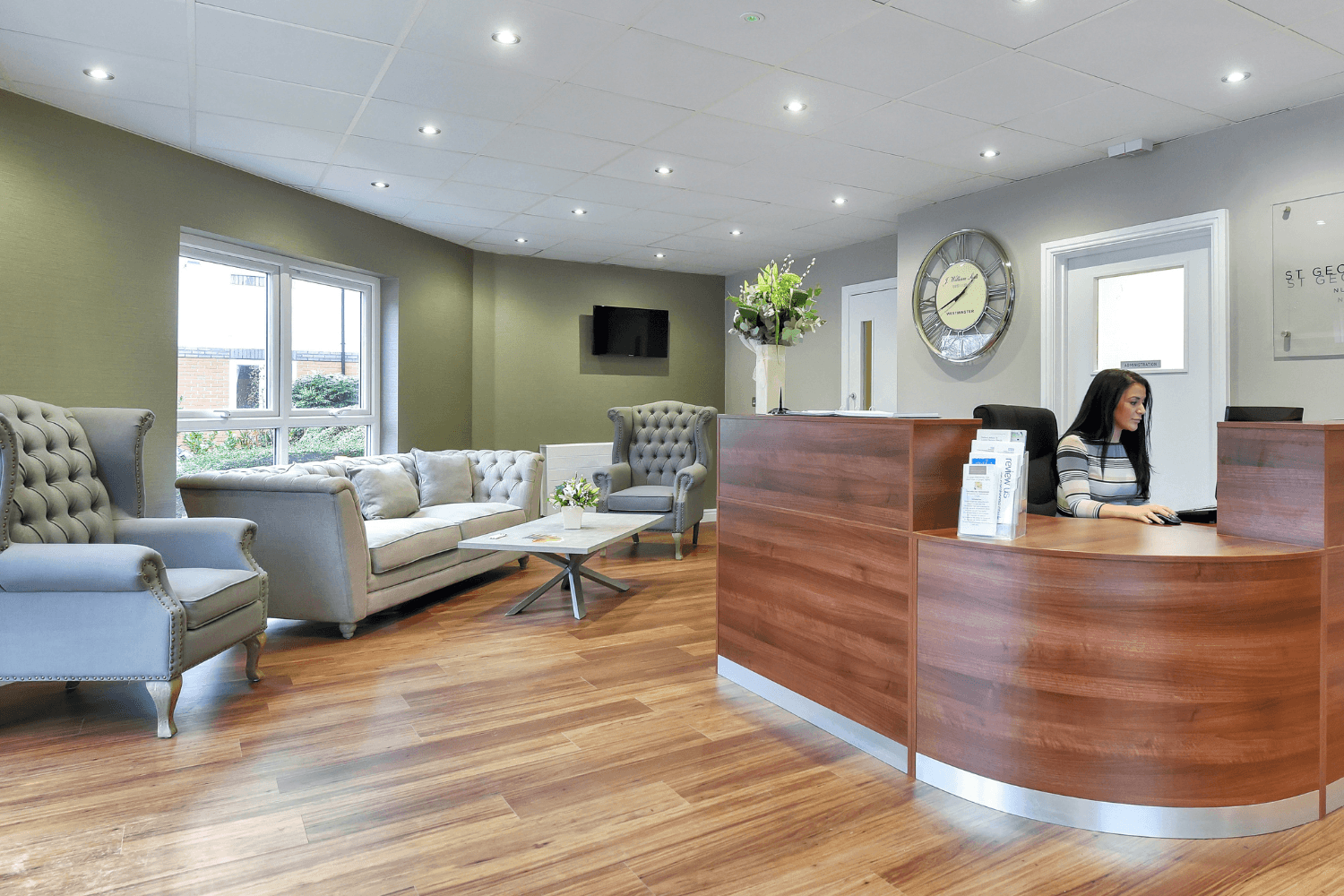 A modern reception area with a wooden desk, grey armchairs, a sofa, and a wall-mounted television