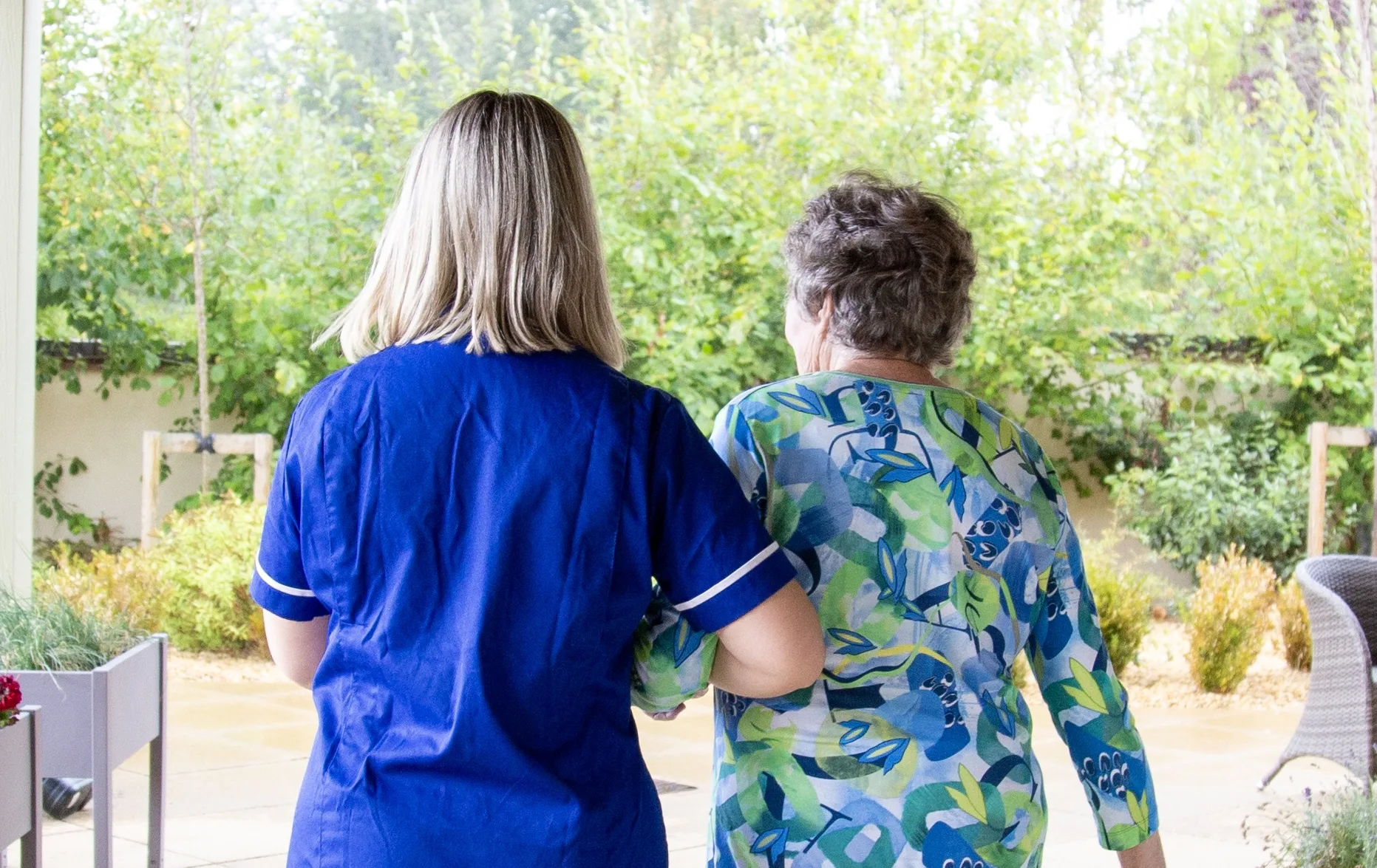A carer walking arm in arm with an older woman across a garden terrace