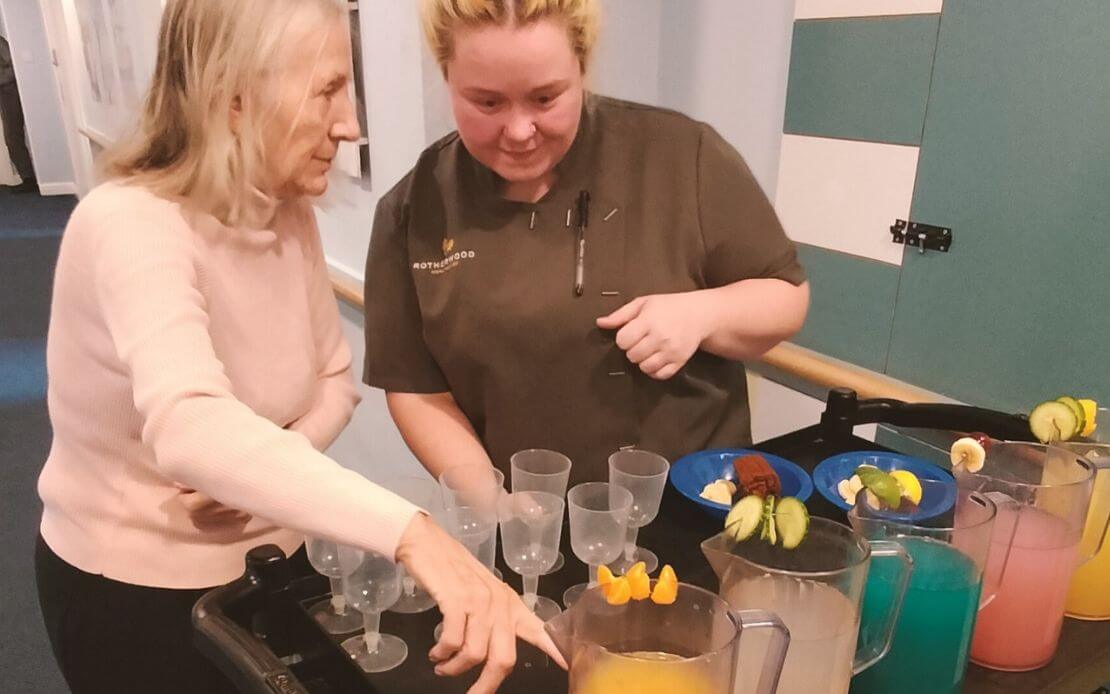 Lifestyle coordinator and older lady selecting a jug of juice from a beverage trolley filled with various colourful options