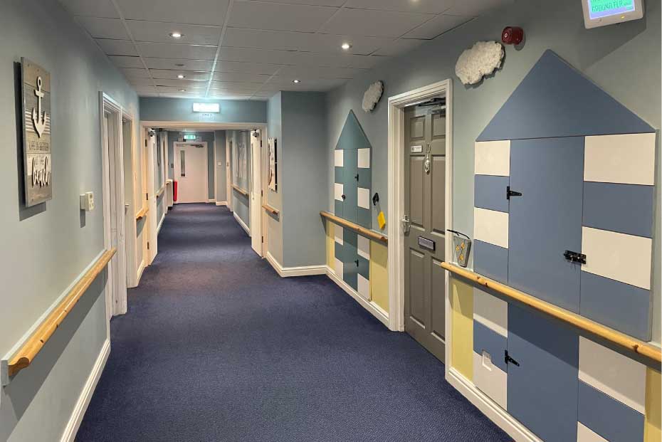 A corridor with blue carpet, walls painted and decorated to resemble blue beach huts, multiple doors on either side, and a wooden handrail