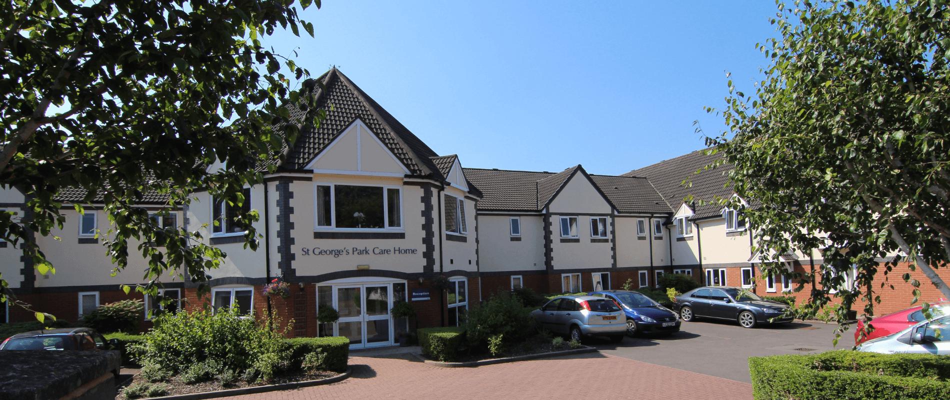 St George's Park front entrance and car park with several cars parked. The two-story building has white walls with dark roofing, and multiple windows.