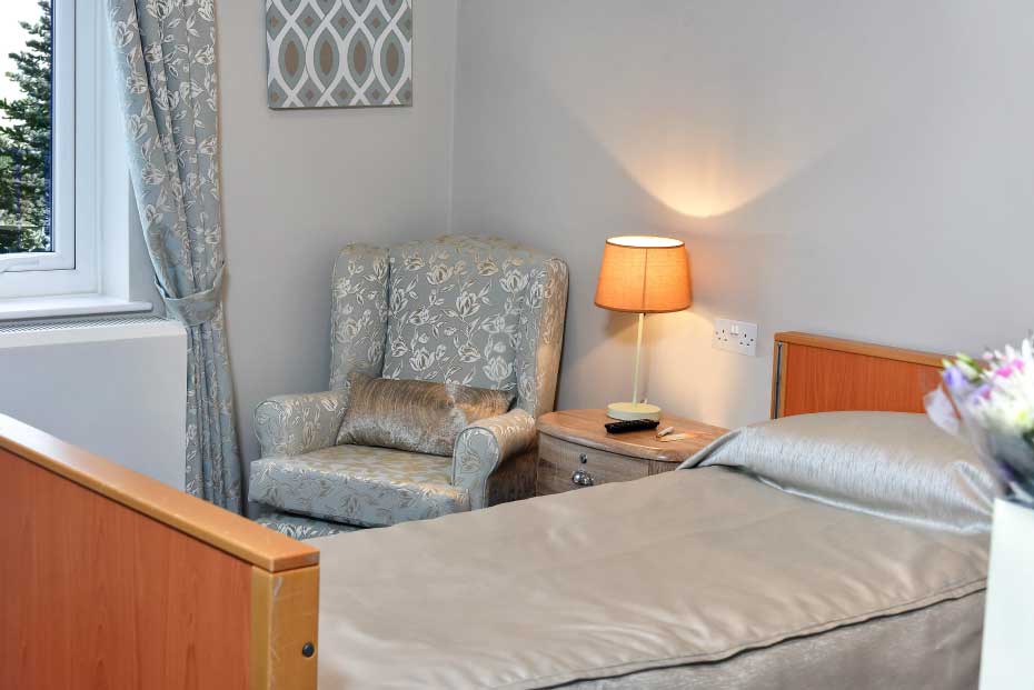 A cosy bedroom with an upholstered armchair, a lit table lamp, and a neatly made bed with a silver comforter.