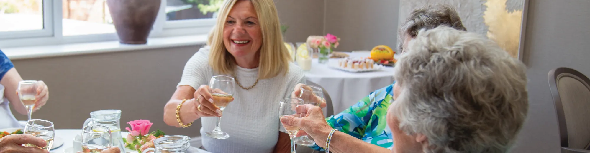 A smiling woman in a white top raising a glass of wine with older women around a dining table