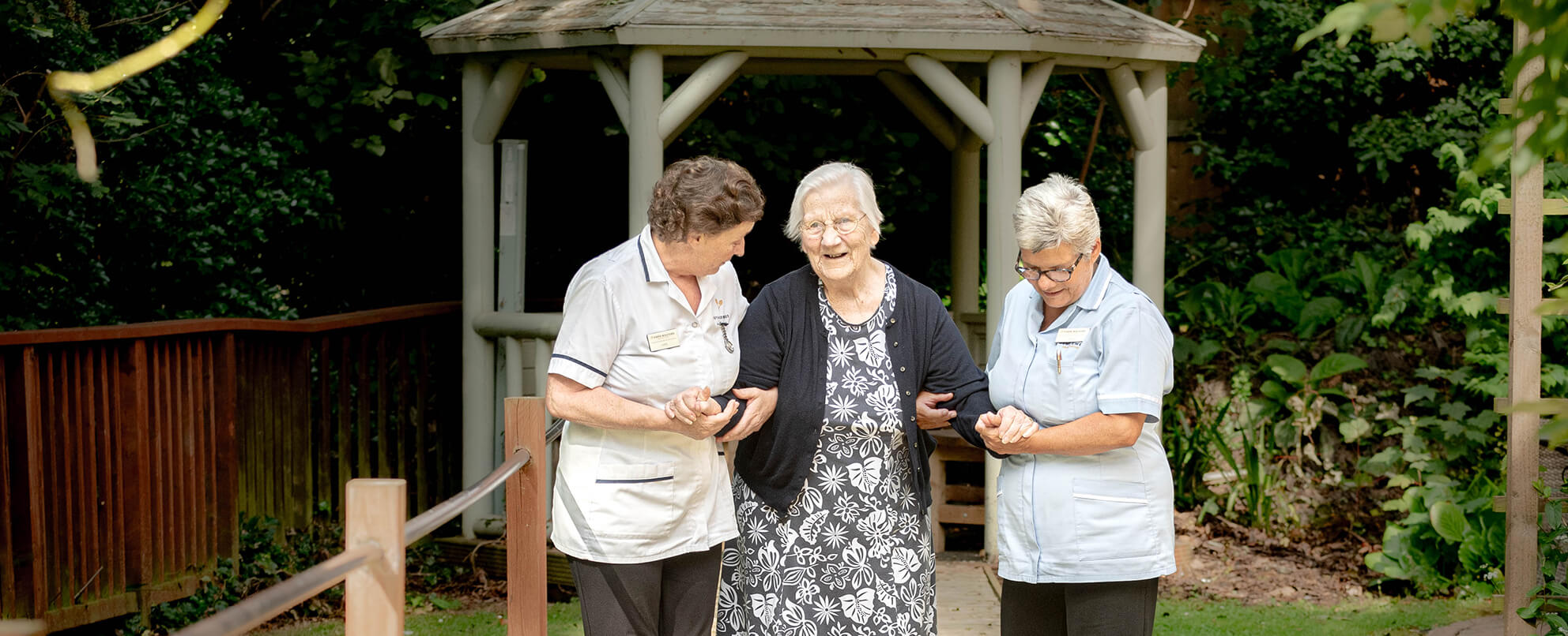 Two healthcare workers assist an older lady walking in a garden.