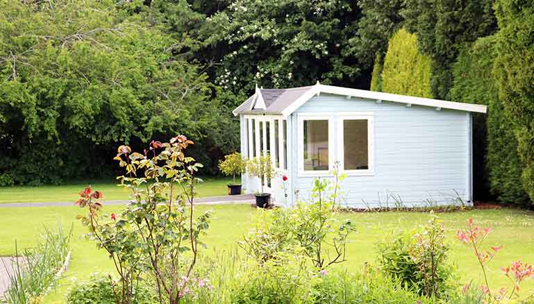 Quaint light-blue summer house with large windows, situated in a landscaped garden and lawn. The summer house is surrounded by an array of greenery, including trees and a blooming rose bush to the left.