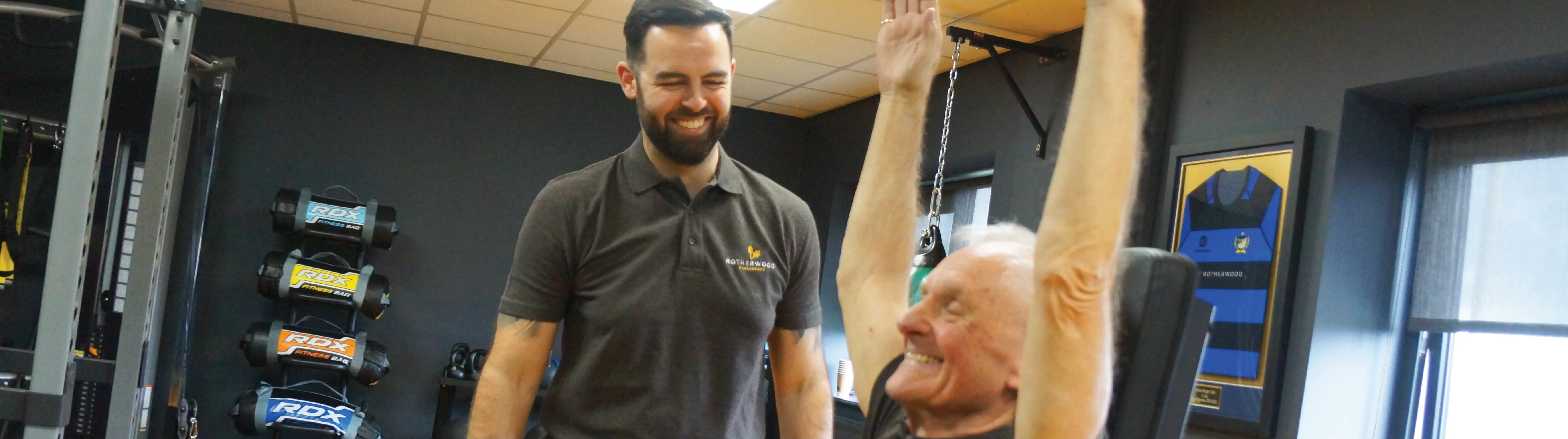 An elderly man smiling whilst exercising on gym equipment with a personal trainer